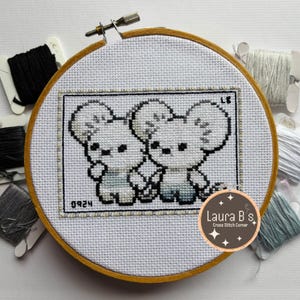 May include: A cross-stitch embroidery hoop featuring two cartoon mice. The mice are white with gray accents and are holding hands. The embroidery is framed in a rectangular border. The hoop is made of wood and is surrounded by spools of thread.