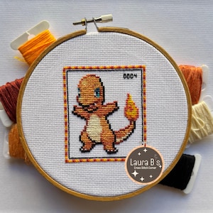 Peut inclure: Un motif de point de croix représentant un personnage de dessin animé orange et jaune de Pokemon, Charmander, sur un tissu blanc dans un cerceau de broderie en bois. Le cerceau est entouré de fils à broder orange, rouge, blanc et noir.