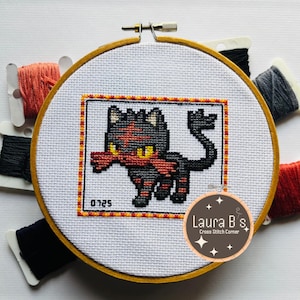 May include: A cross stitch pattern of a black and red Pokemon character, with a yellow scarf, in a wooden embroidery hoop. The pattern is surrounded by a yellow and orange border. The hoop is surrounded by colourful embroidery floss.