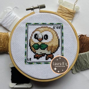 May include: A cross stitch pattern of a brown and white owl with green eyes and a green bow tie. The owl is surrounded by a green and white border. The pattern is on a white fabric and is in a wooden embroidery hoop. The text "Laura B's Cross Stitch Corner" is on a brown circle with white stars.