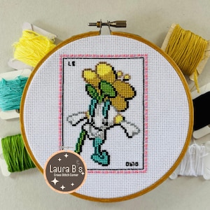 May include: A cross stitch embroidery hoop with a yellow flower design. The hoop is surrounded by colorful embroidery floss spools. The design includes the text "Laura B's Cross Stitch Corner" and "0510".