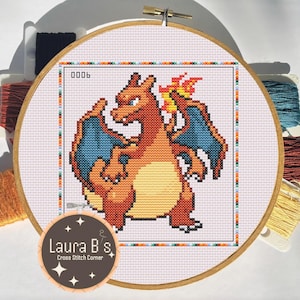 May include: A round wooden embroidery hoop containing a cross-stitch pattern of a pixelated orange dragon-like creature with blue wings and a flame on its tail. The text 0006 is visible in the corner, surrounded by colorful embroidery floss bobbins.