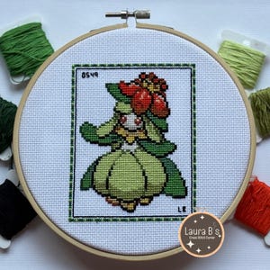 Op de afbeelding: Kruissteekpatroon van een groen en rood Pokemon-personage in een hoepel. Het patroon is gelabeld "0549" en "LE". De hoepel is omgeven door borduurgaren in verschillende kleuren.
