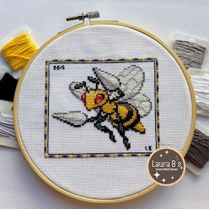Puede incluir: Un bordado de punto de cruz de una abeja en un aro de madera. La abeja es amarilla y negra con alas blancas, dentro de un borde negro, verde y morado. El texto "0015" está en la esquina superior izquierda. Hilo de bordar de varios colores rodea el aro.