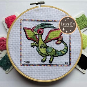 Puede incluir: Un bordado de punto de cruz de una criatura verde y amarilla con forma de dragón, con ojos y alas rojas, en un bastidor de bordado de madera. El diseño incluye el número "0330" y las iniciales "LK". El fondo incluye madejas de hilo de bordar de colores.