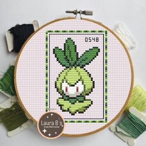 May include: Cross-stitch embroidery of a green cartoon character with red eyes, framed in a wooden hoop. The design includes the number 0548. The embroidery is surrounded by green and black thread spools and white floss.