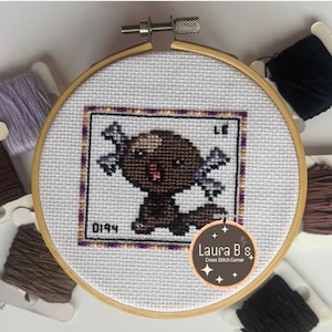 May include: A cross-stitch embroidery of a cartoon axolotl in a wooden hoop. The axolotl is brown with purple accents, framed by a purple and black border. The text "0194" is visible, along with the maker's logo.