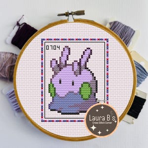 May include: A cross-stitch design of a purple creature with green accents, framed in a wooden hoop. The design includes the number "0704" and is surrounded by red and blue stitches. The hoop is set against a white background with spools of thread.