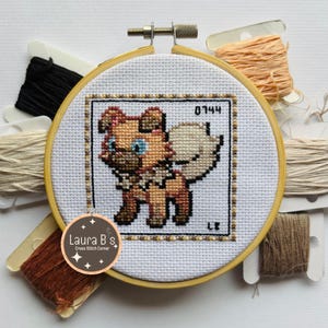 Könnte beinhalten: Ein Kreuzstichmuster eines braunen und weißen Hundes mit einem blauen Auge. Das Muster befindet sich in einem runden Holzstickrahmen mit beigefarbenem Hintergrund. Das Muster ist mit der Nummer 0144 und den Buchstaben LE gekennzeichnet.