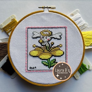 May include: A cross stitch pattern of a yellow flower with a white fairy in the center. The pattern is framed in a wooden hoop and surrounded by embroidery floss in various colors. The text "Laura B's Cross Stitch Corner" is visible in the lower right corner.