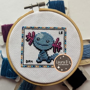 May include: Cross-stitch embroidery of a blue cartoon creature with pink accents, framed in a wooden hoop. The design includes the number "0194" and the initials "LB". The embroidery is surrounded by spools of colorful thread.