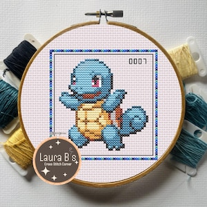 May include: Cross-stitch embroidery of a blue Squirtle character from the Pokémon series, framed in a wooden hoop. The design features a light blue Squirtle with a tan shell, pink eyes, and the number "0007". The hoop is surrounded by spools of thread.