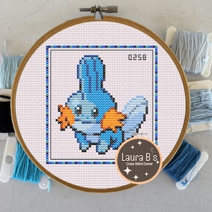 Pode incluir: Bordado em ponto cruz de uma criatura azul de desenho animado com detalhes em laranja, emoldurado em uma borda quadrada. O bordado está em um bastidor de madeira, com o número "0258" visível. O logotipo do artista, "Laura B's Cross Stitch Corner", é visível.