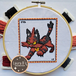 May include: A cross-stitch embroidery hoop featuring a red and black Pokemon character, "0126", and "Laura B's Cross Stitch Corner" on a brown tag.
