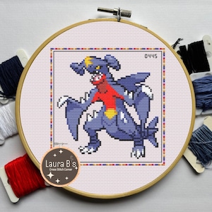 Puede incluir: Bordado de punto de cruz de un personaje Garchomp en un aro de madera. El diseño presenta una criatura azul, roja y amarilla con el número 0445. El aro está rodeado de carretes de hilo de varios colores, incluyendo rojo, blanco y azul.