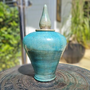May include: A teal blue ceramic vase with a pointed lid. The vase is sitting on a dark brown circular surface with intricate designs.