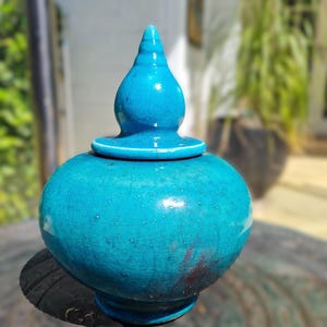 May include: A teal blue ceramic jar with a lid. The jar has a rounded body and a pointed lid. The jar is sitting on a wooden surface.