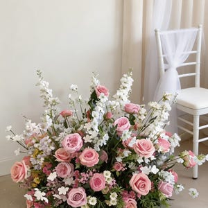 May include: A large floral display of pink roses and white flowers. The arrangement is on the floor, with a white chair and sheer curtain in the background. The flowers are in full bloom, creating a soft, romantic aesthetic, suitable for weddings.