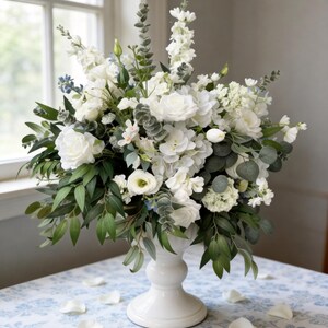 May include: A large, elegant floral arrangement in a white pedestal vase. The bouquet features white roses, hydrangeas, and other flowers, with green eucalyptus and other foliage. White petals are scattered on the blue and white patterned tablecloth.