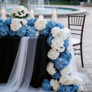 Op de afbeelding: Een tafel versierd voor een buitenevenement met blauwe en witte hortensia's, kaarsen in glazen cilinders en een zwart tafelkleed. Een witte, doorschijnende stof drapeert over de rand van de tafel. Een fontein is zichtbaar op de achtergrond.
