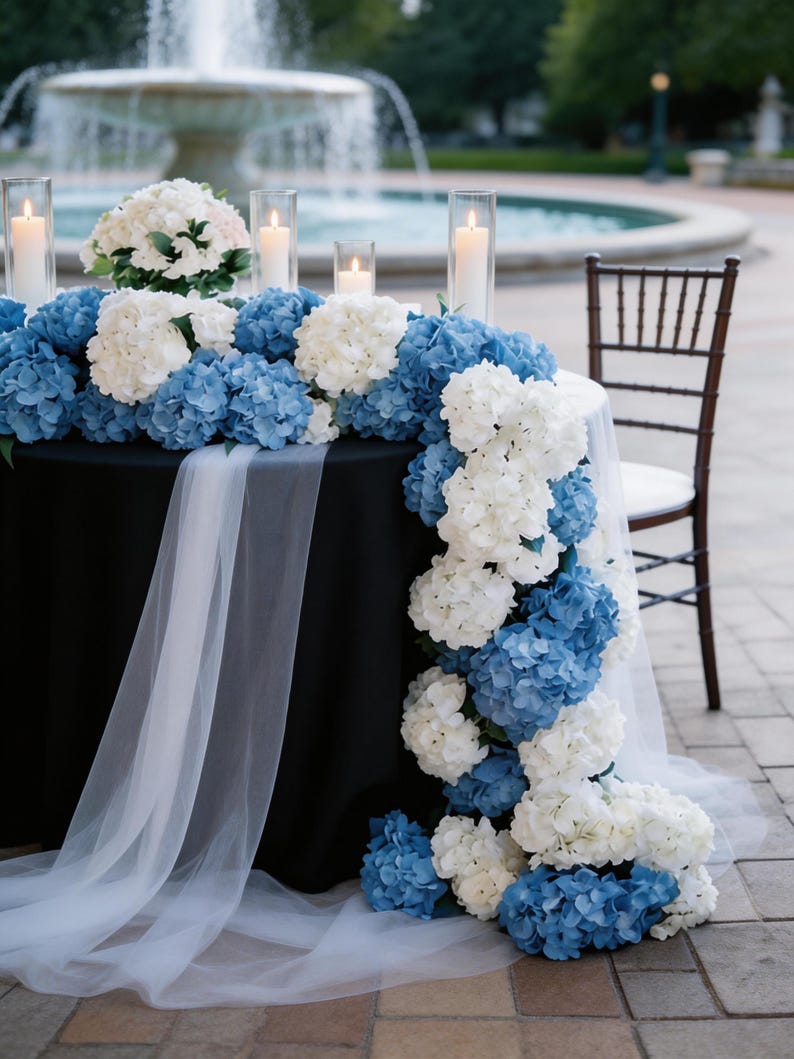Op de afbeelding: Een tafel versierd voor een buitenevenement met blauwe en witte hortensia's, kaarsen in glazen cilinders en een zwart tafelkleed. Een witte, doorschijnende stof drapeert over de rand van de tafel. Een fontein is zichtbaar op de achtergrond.