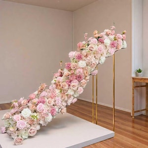 May include: A display of pink, white, and peach roses and other flowers cascading from a gold metal frame. The floral arrangement sits on a white platform. The background is a neutral beige, with wooden flooring.