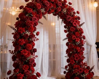 May include: A large, arch-shaped floral arrangement composed of various shades of red roses and other flowers. The arch is set against a backdrop of sheer white curtains and a polished wooden floor, creating an elegant display.