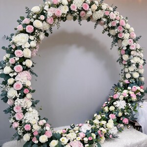 May include: A large circular floral arch arrangement featuring a dense composition of white and light pink roses, white hydrangeas, and green eucalyptus foliage. The flowers are arranged in a continuous ring shape with varied textures.