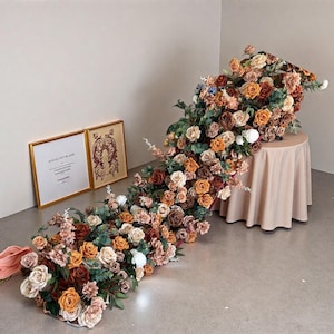 May include: A floral display of orange, brown, and cream roses cascades from a small round table. The arrangement includes greenery and is set against a neutral background. Two framed artworks are visible.