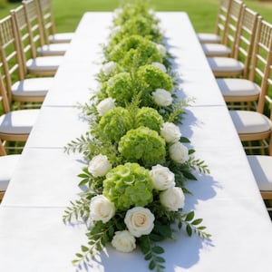 Op de afbeelding: Een lange witte tafel is versierd met een bloemstuk van groene hortensia's, witte rozen en groen. Gouden stoelen met witte kussens zijn aan weerszijden van de tafel opgesteld, buiten op een gazon.