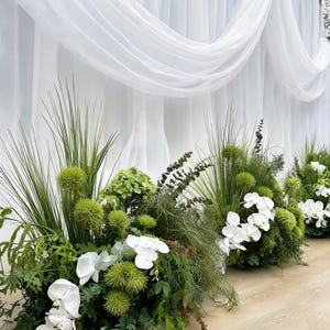 以下が含まれることがあります： 白い蘭、鮮やかな緑のダイアンサス、背の高い草の葉を組み合わせたフラワーアレンジメント。柔らかなドレープを描く白い布の背景の前に配置されており、重なり合うような優雅で洗練された外観を作り出しています。