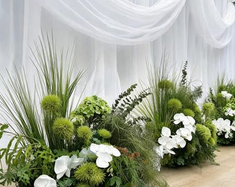 Op de afbeelding: Een bloemstuk met witte orchideeën, felgroene duizendschoon en lange grassprieten. Het arrangement staat voor een witte, gedrapeerde stoffen achtergrond met zachte, vloeiende plooien die zorgen voor een gelaagde en elegante uitstraling.