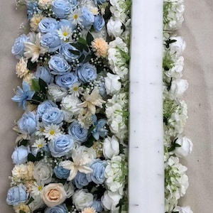 以下が含まれることがあります： ライトブルー、ホワイト、ペールピーチの造花のバラとデイジーを組み合わせたフラワーアレンジメント。中央を縦に長い長方形の白いフォームベースが貫いており、ピンで花の茎が固定されているのが見えます。
