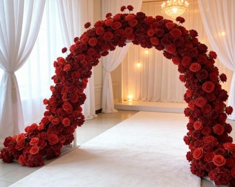 May include: A large, arch-shaped floral arrangement made of red roses. The arch is supported by a white frame and sits on a white carpet. The background includes white curtains, a chandelier, and soft lighting, creating an elegant setting for a wedding or event.