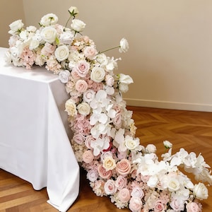 May include: A cascading floral arrangement featuring white, pink, and blush roses, orchids, and hydrangeas. The flowers spill from a white tablecloth onto a wooden floor, creating a romantic and elegant display. Perfect for weddings or special events.