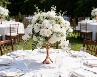 May include: A beautifully arranged table setting for an outdoor event. The centerpiece is a large bouquet of white roses and greenery in a copper-colored vase. The table is set with white plates, napkins, silverware, and wine glasses. Wooden chairs surround the table.
