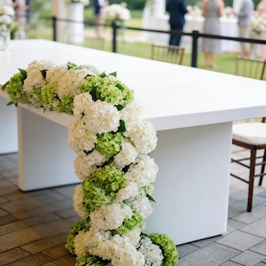 Op de afbeelding: Een witte rechthoekige tafel versierd met trapsgewijze witte en groene hortensia bloemen. De bloemen zijn langs de rand en de zijkant van de tafel gerangschikt, waardoor een decoratieve display voor een evenement ontstaat.