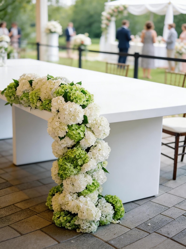 Op de afbeelding: Een witte rechthoekige tafel versierd met trapsgewijze witte en groene hortensia bloemen. De bloemen zijn langs de rand en de zijkant van de tafel gerangschikt, waardoor een decoratieve display voor een evenement ontstaat.