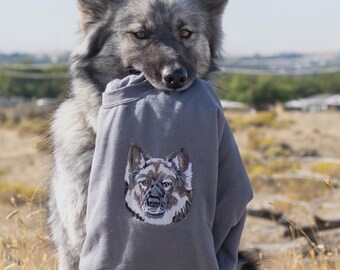 May include: A gray wolfdog wearing a gray sweatshirt with a wolf embroidered on the front. The sweatshirt has the text "CORGO ART STUDIO" on the bottom.