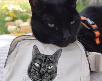 Puede incluir: Un gato negro con ojos verdes descansa sobre un bolso cruzado color crema. El bolso presenta un bordado de la cara de un gato. El gato lleva un arnés naranja. El fondo incluye una zona de césped y un árbol.