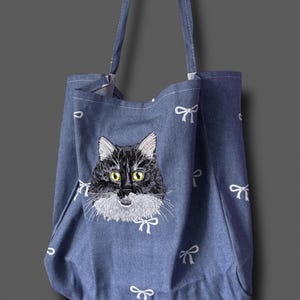 May include: Blue denim tote bag with an embroidered black and white cat face and yellow eyes. The bag has white bow accents and long straps. A stylish accessory for carrying items, perfect for cat lovers.