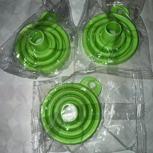 May include: Three green, collapsible funnels in clear plastic packaging. Each funnel has a circular design with concentric rings and a small loop for hanging. The funnels are made of a flexible material.