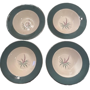 May include: A set of four round ceramic bowls, each featuring a wide teal rim and a cream-coloured centre. A small, delicate floral motif with purple buds and green leaves is painted in the middle of each bowl, providing a consistent design.