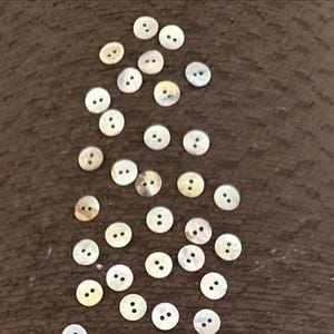 Op de afbeelding: Een verzameling kleine, ronde, iriserende parelmoeren knopen, elk voorzien van twee centrale gaten. Deze ronde sluitingen vertonen een natuurlijke, parelmoerachtige glans met subtiele variaties in witte en crèmetinten over hun gladde oppervlakken.