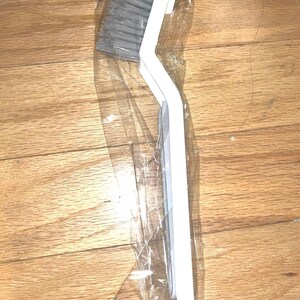 May include: A white handled cleaning brush with gray bristles, packaged in clear plastic. The brush has a curved design and is likely for cleaning hard-to-reach areas. The bristles appear to be tightly packed.