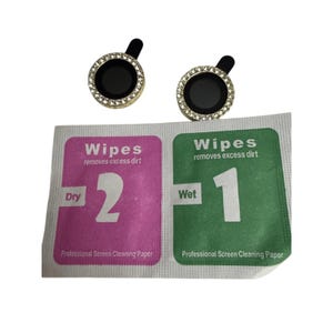 Puede incluir: Dos protectores de lente de cámara negros y dorados con un borde de diamantes de imitación. Debajo hay dos toallitas de limpieza, una verde y otra rosa, con el texto "Wipes removes excess dirt" y los números 1 y 2.