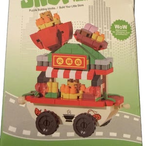 Puede incluir: Una caja de bloques de construcción con un modelo de puesto de comida colorido con detalles en rojo, verde y blanco. El empaque muestra el texto SHOP vendor, Puzzle Buliding blocks.. / Build Your Little Store y caracteres chinos.