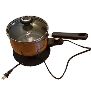 Può includere: Un pentolino elettrico con esterno a venature di legno, manico nero e coperchio in vetro trasparente con pomello nero. La base è nera e include una manopola di controllo della temperatura integrata e un cavo di alimentazione nero.