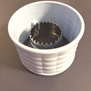 Könnte beinhalten: Ein weißer Plastik-Ausstecher mit geriffeltem Rand und einem kleineren, gezackten Metallausstecher im Inneren. Die Ausstecher sind zum Ausstechen von Keksen oder anderen Backwaren gedacht.