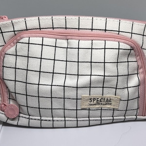 Könnte beinhalten: Ein weißes Federmäppchen aus Segeltuch mit schwarzem Karomuster, ausgestattet mit einem rosa Reißverschluss und einem rosa Zipper. Ein rechteckiger beiger Stoffaufnäher auf der Vorderseite zeigt den Text "SPECIAL" und "YOU ARE ONE IN A MILLION".